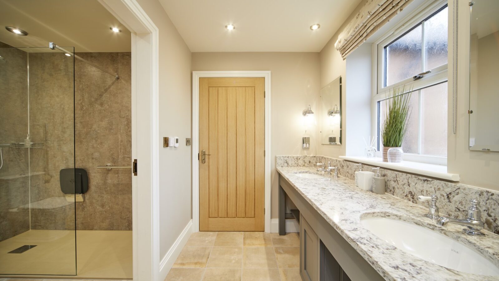 The Stables offers a modern bathroom featuring a granite countertop, double sink, and large mirror. A walk-in shower sits elegantly beside a wooden door, all bathed in natural light from the window.