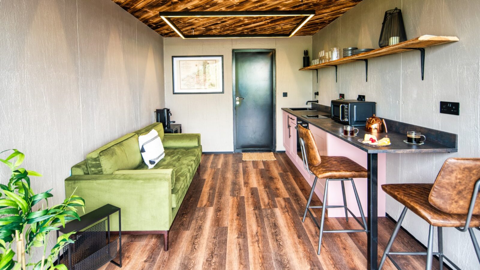 A cozy modern room with a green sofa, barstools at a kitchen counter, and wooden accents on the ceiling and floor evokes the charm of a rustic boathouse.