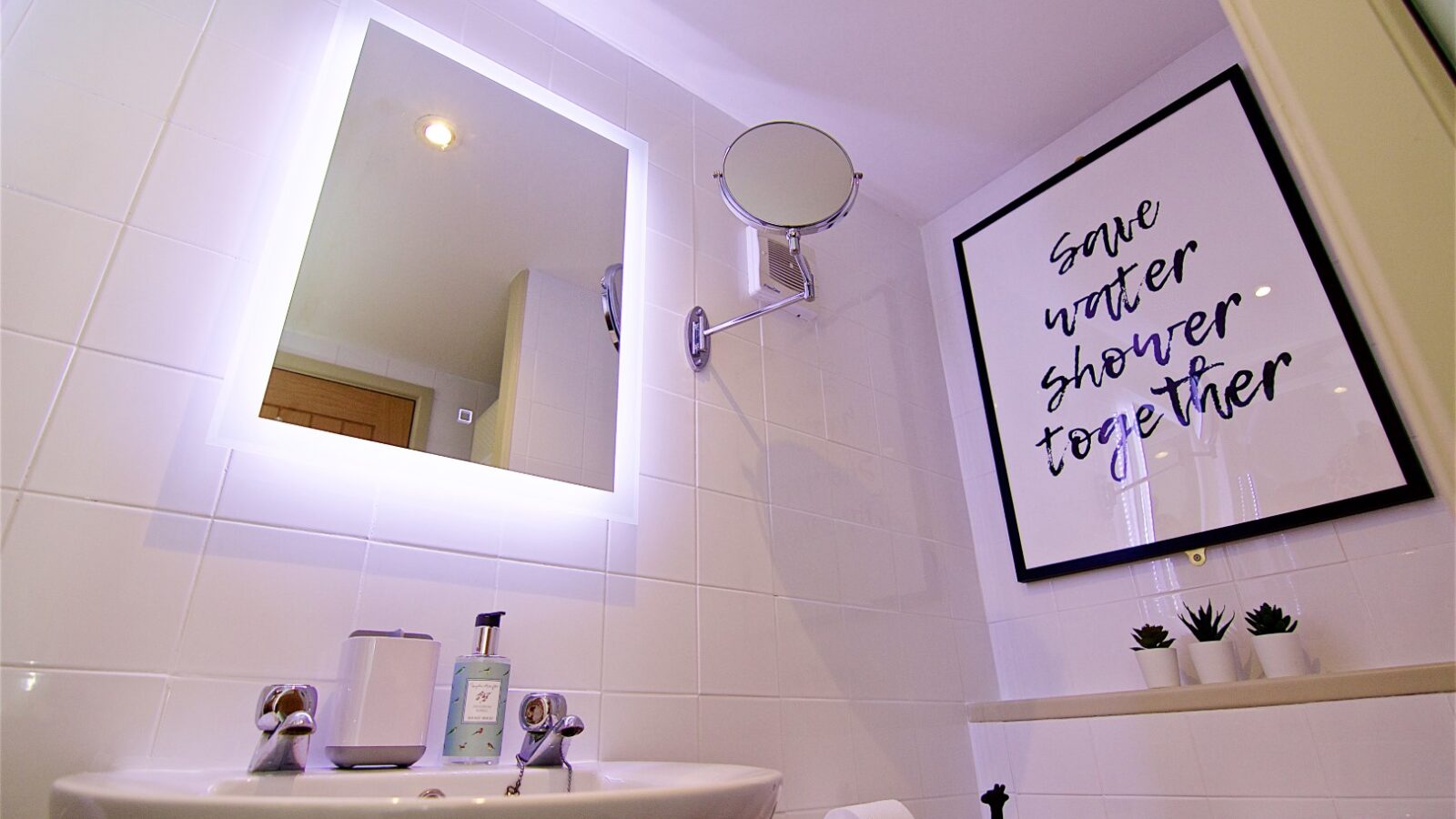 The bathroom at Georges Getaway features an illuminated mirror, a framed quote about saving water, a sleek sink with a soap dispenser, and small plants on the shelf adding a touch of nature.