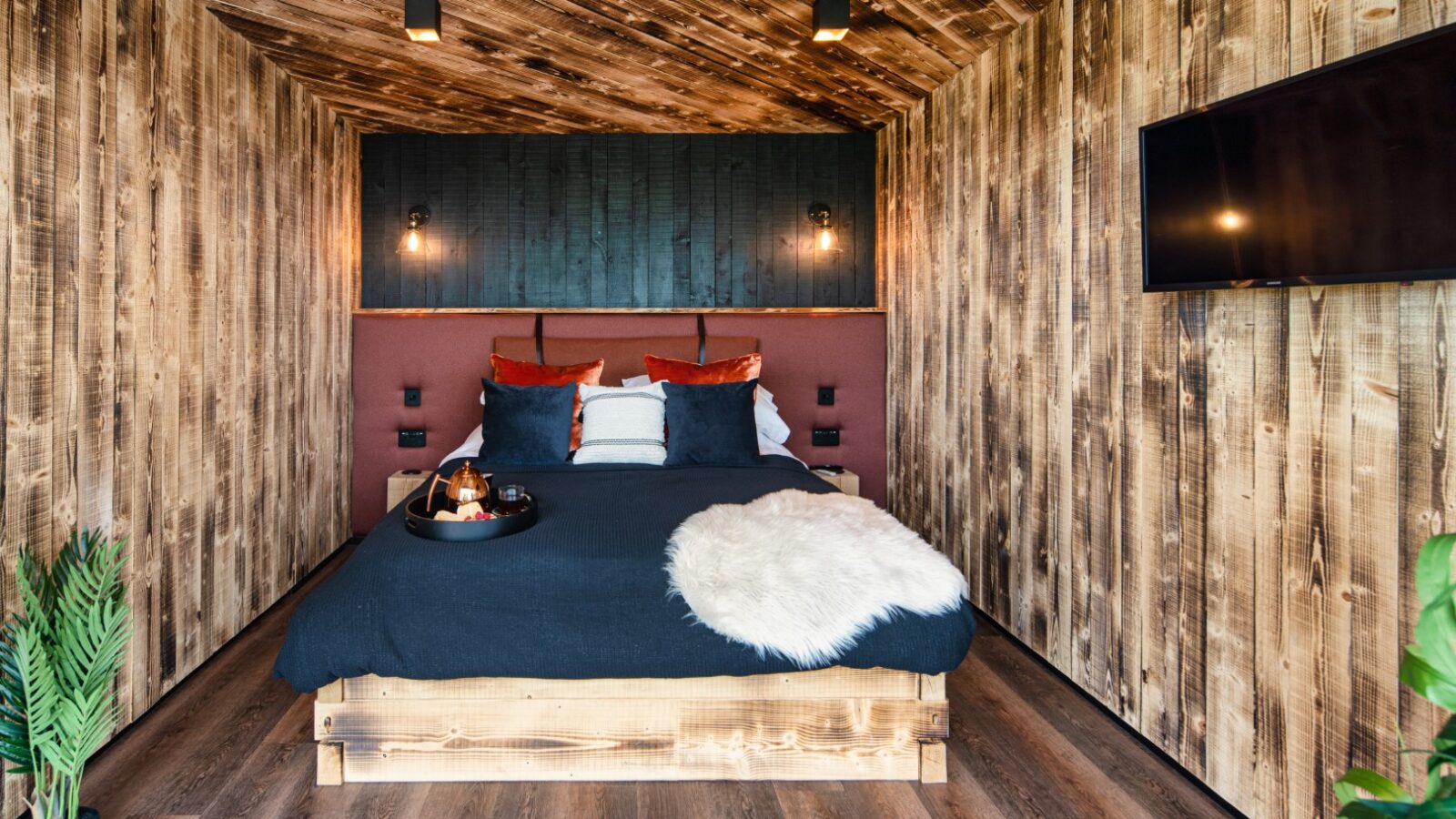 A cozy, wood-paneled bedroom reminiscent of the warmth found at the Rusty Boathouse, featuring a neatly made bed with decorative pillows and a wall-mounted TV on the right.