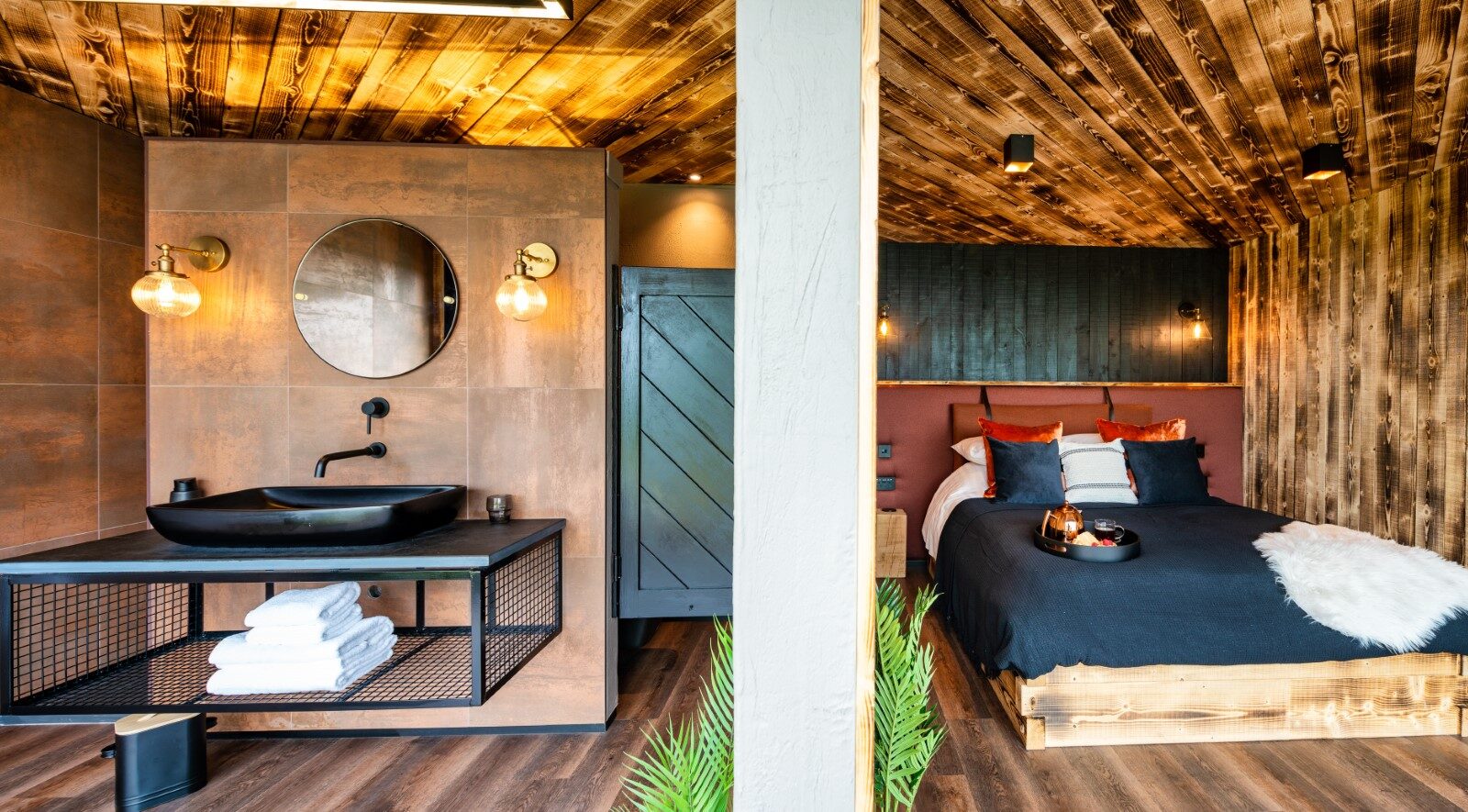 The modern interior exudes a rustic charm reminiscent of a rusty boathouse. It features a wood-paneled bedroom and a stylish bathroom with a round mirror, black sink, and warm lighting.