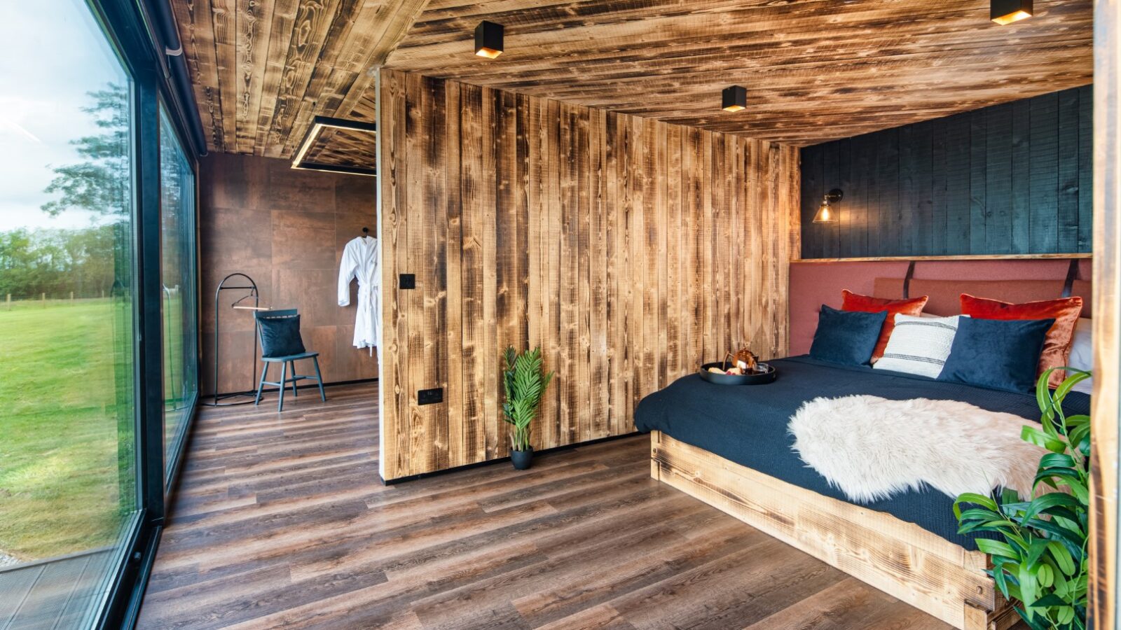 Modern bedroom with wood paneling reminiscent of a rustic boathouse, featuring a large window and a neatly made bed. A chair and robe add to the serene atmosphere in the background.