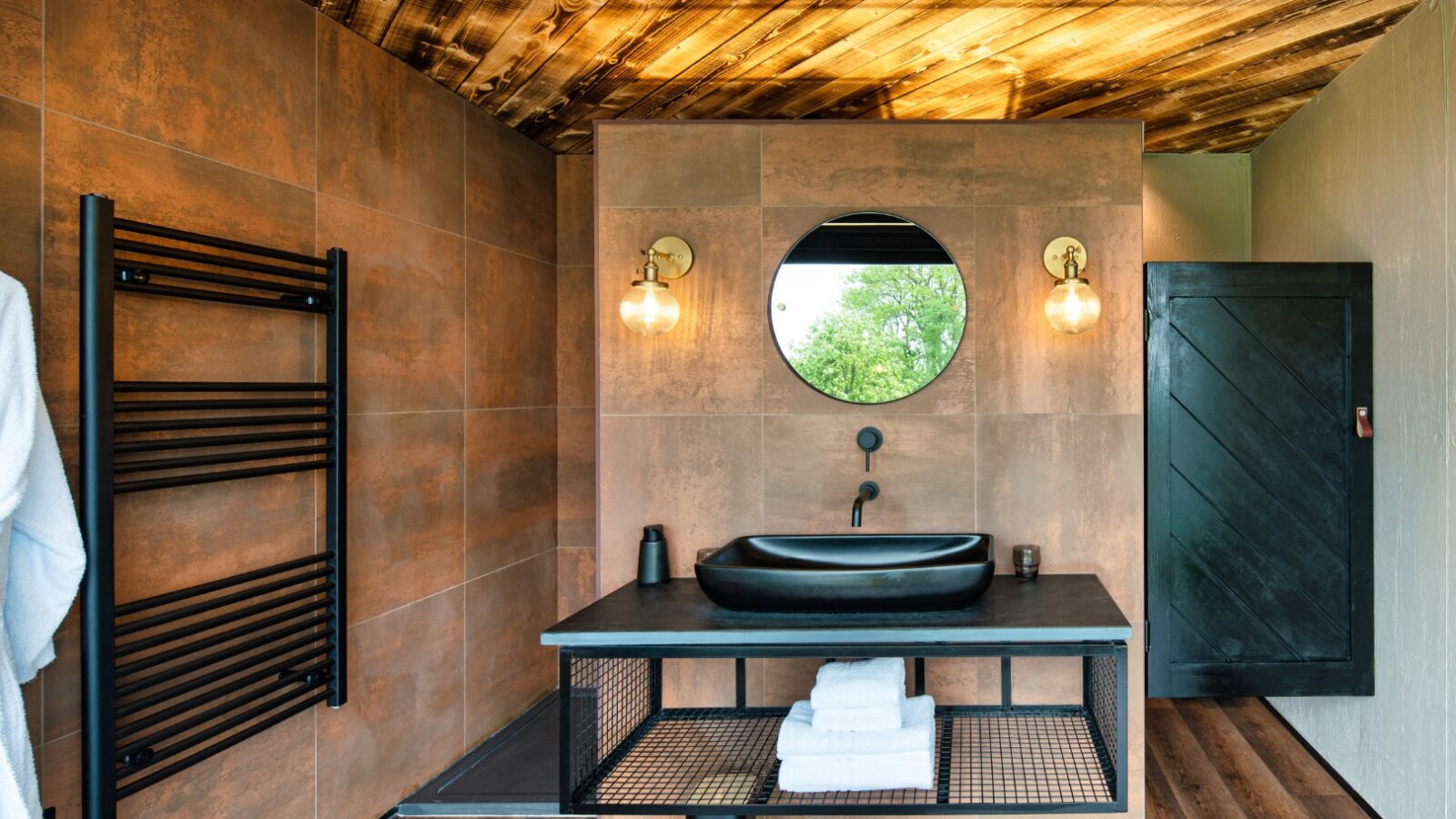 This modern bathroom, echoing a rustic boathouse vibe, features wood accents and includes a round mirror, black sink, wall-mounted lights, and a stylish black towel rack.