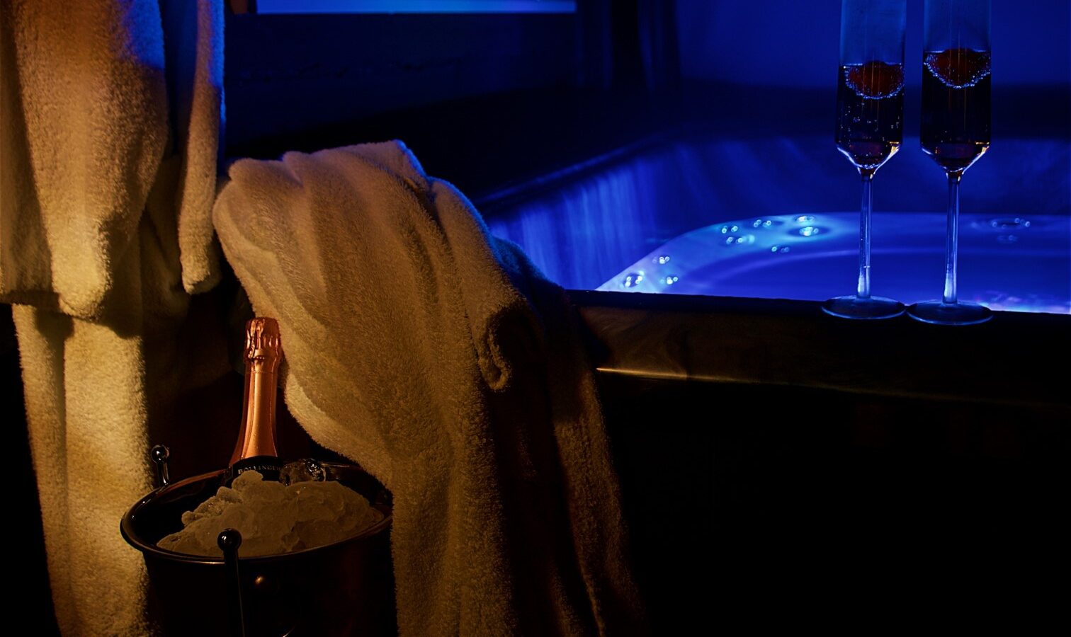 Two champagne glasses perched on the glowing edge of the hot tub, surrounded by tranquility at Georges Getaway, with a towel draped nearby and a bottle of bubbly nestled in an ice bucket.