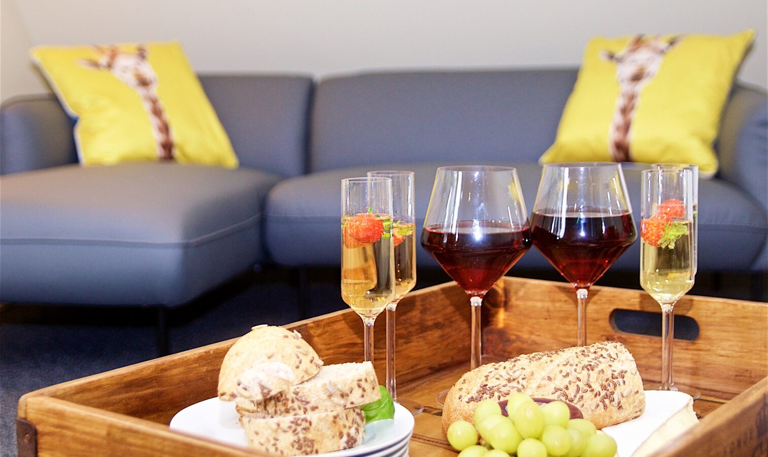 Wooden tray with wine glasses, bread, and grapes on a table in front of a sofa adorned with giraffe-themed pillows, capturing the essence of a perfect Georges Getaway.