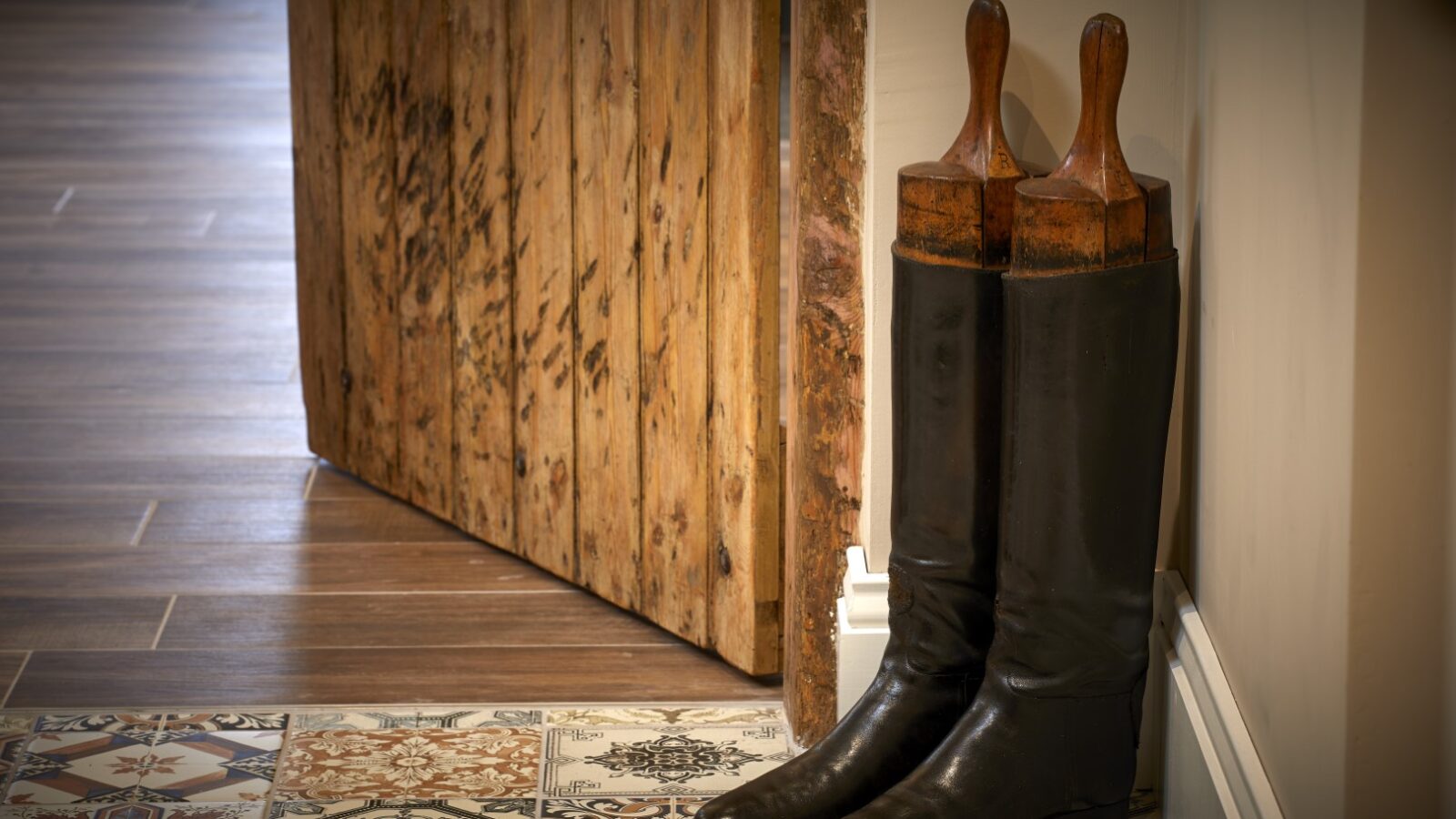 A pair of black leather boots, reminiscent of ones worn in stables, stands with wooden boot trees next to a wooden door on a tiled floor with a colorful pattern.