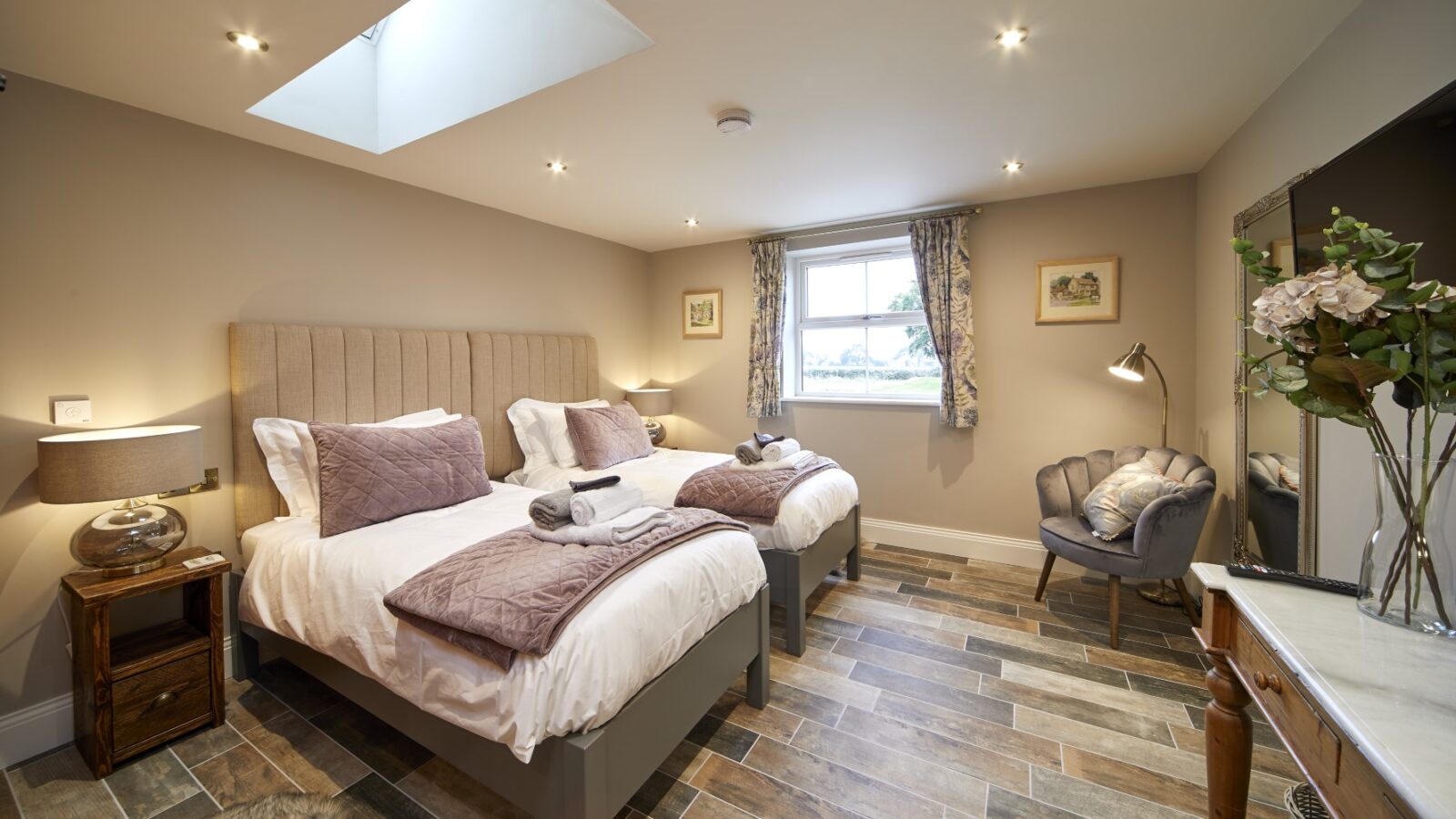 A well-lit bedroom with two single beds, bedside tables, an armchair, a skylight, and a window with patterned curtains brings the rustic charm of converted stables indoors.