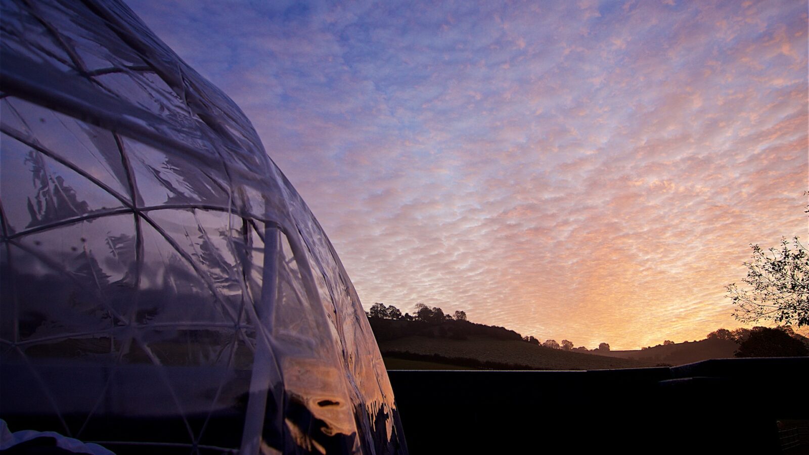 The sunset sky, painted with scattered clouds, casts its glow on the silhouette of the landscape. To the left stands a transparent dome structure, part of Georges Getaway, promising a tranquil retreat amidst nature's beauty.