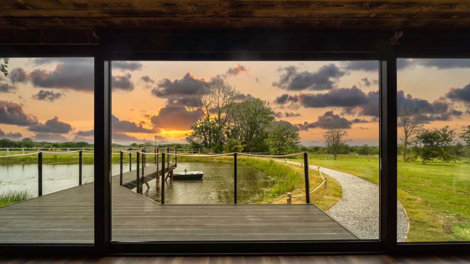 View of a modern deck with a small pond and trees at sunset, seen through a large floor-to-ceiling window, reminiscent of a serene boathouse setting.