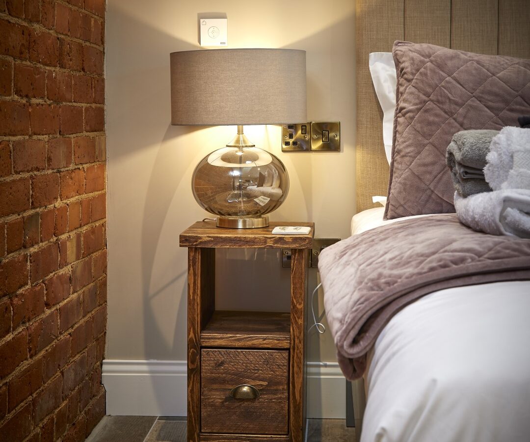 Nestled in The Stables, this cozy bedroom boasts a wooden nightstand, a round glass lamp, and a neatly made bed adorned with quilted pillows and folded towels.