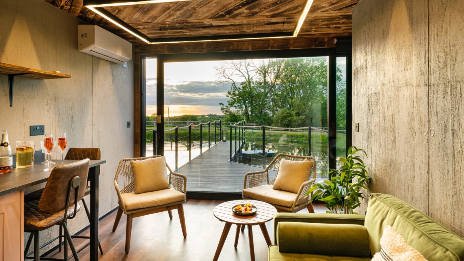 A cozy living room with modern furniture, large windows, and a wooden ceiling offers a view reminiscent of a boathouse deck overlooking a lush landscape.