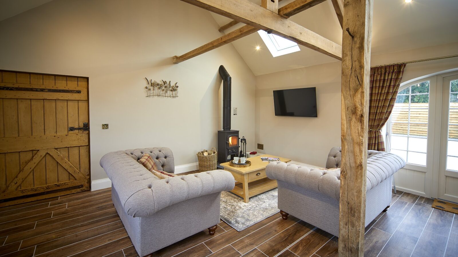 Cozy living room in The Stables features gray sofas, wooden beams, a wood-burning stove, TV on the wall, and wooden flooring.