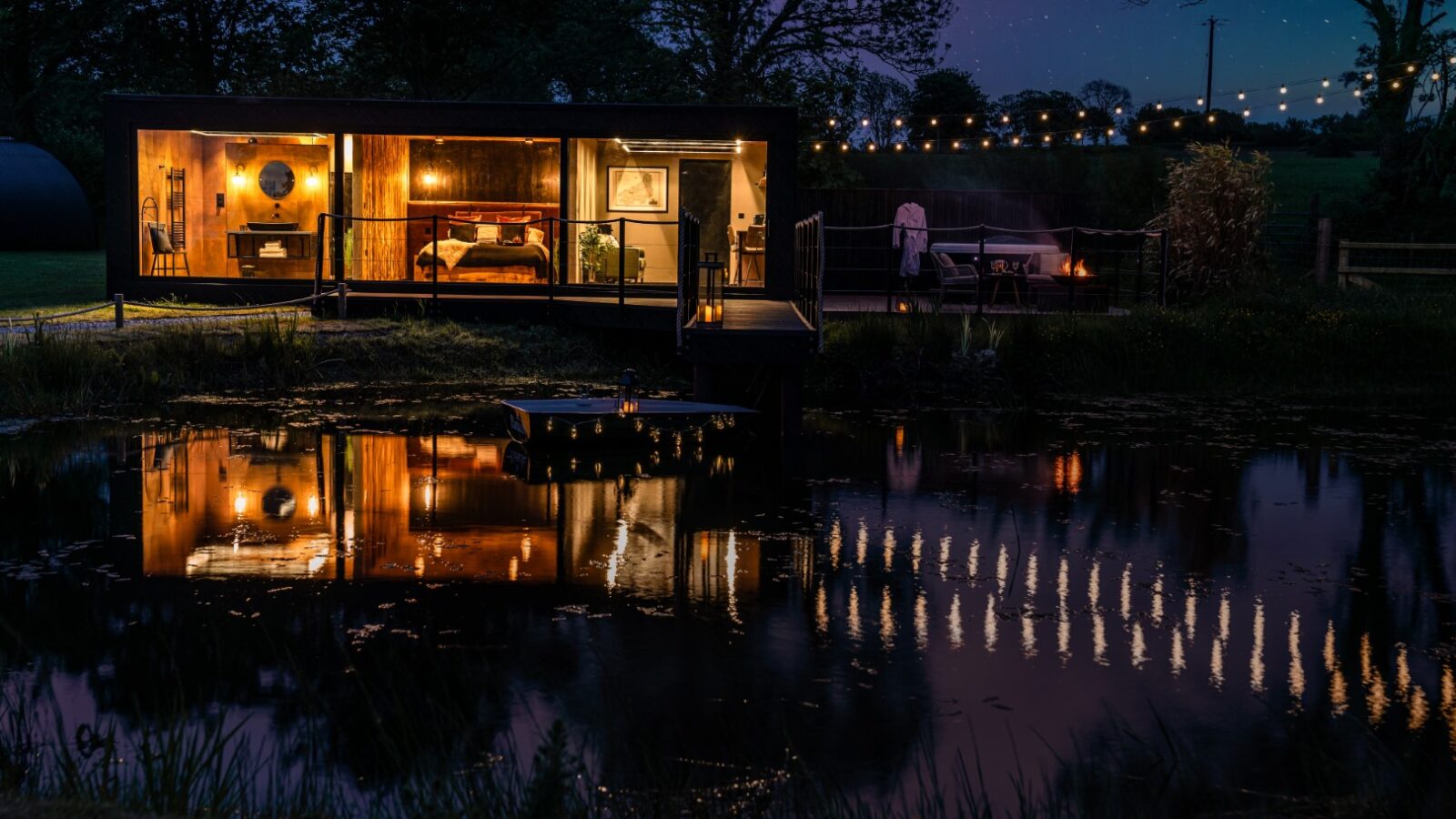 At night, a modern cabin with large windows glows like a rustic boathouse, reflections dancing on the pond. String lights dangle above the outdoor area, creating a warm and inviting ambiance.