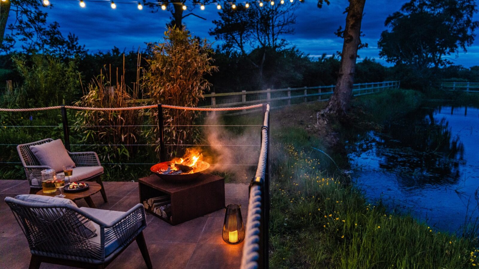 A cozy outdoor patio by a serene pond, reminiscent of a rustic boathouse vibe, features two inviting chairs, a fire pit, a small table, and enchanting overhead string lights that create the perfect evening glow.