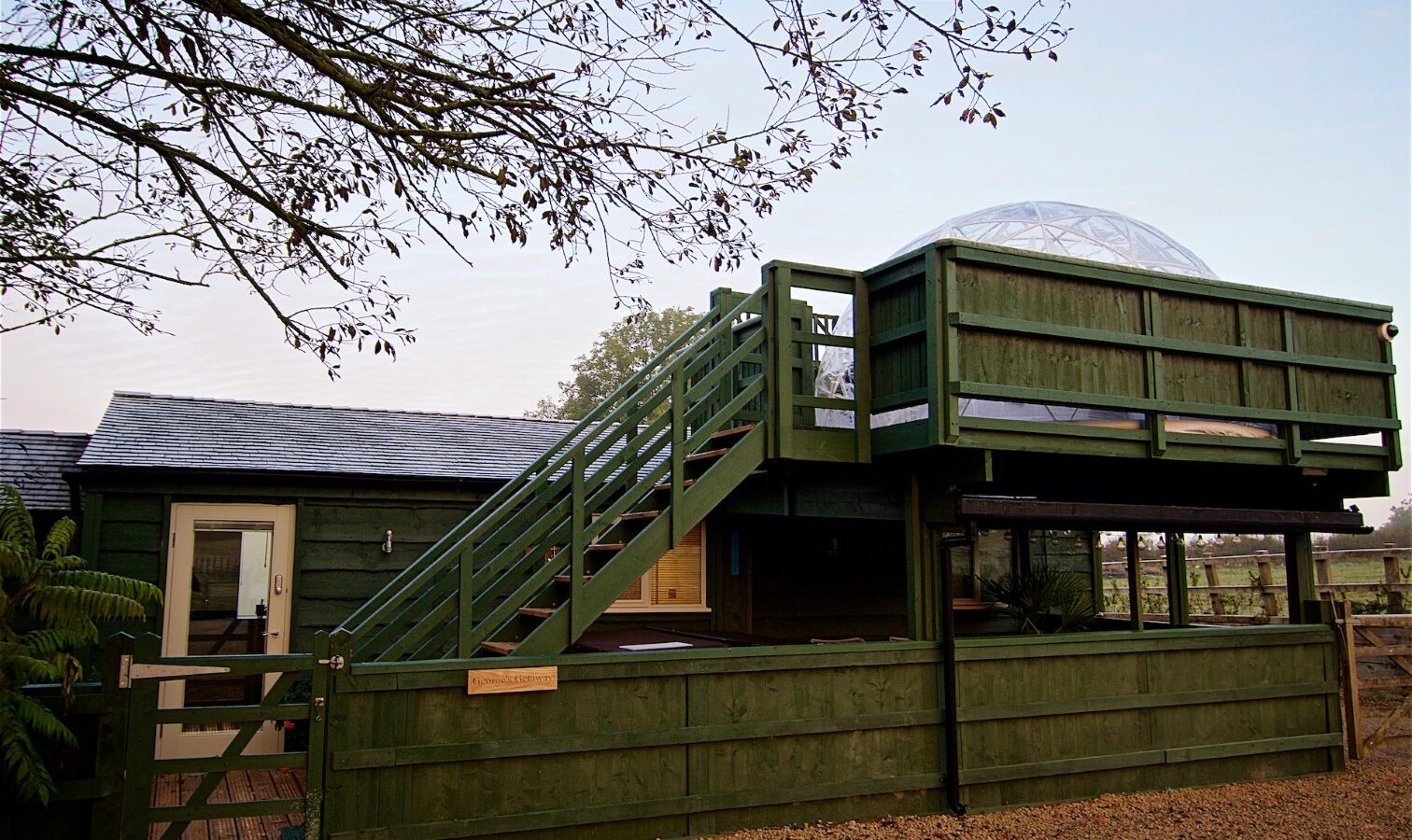 Green wooden structure with a staircase leading to a dome-covered deck, perfect for a serene vacation retreat. A gravel path winds through the travel destination's foreground as tree branches sway gently above.