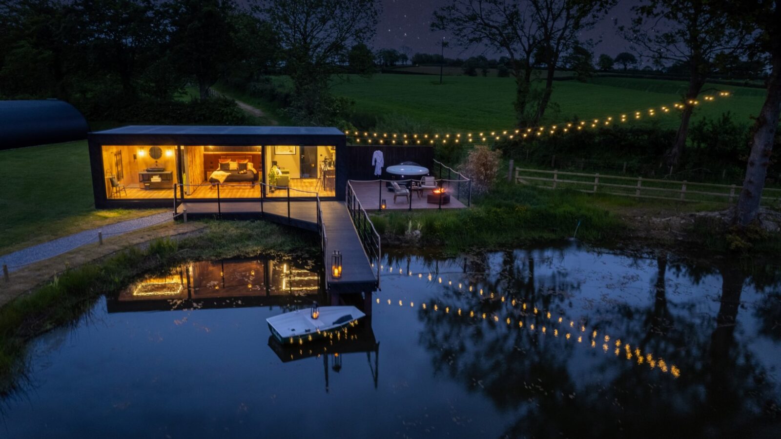 Modern house with large windows by a pond at night, illuminated by string lights reflecting on the water, near a charming rusty boathouse.