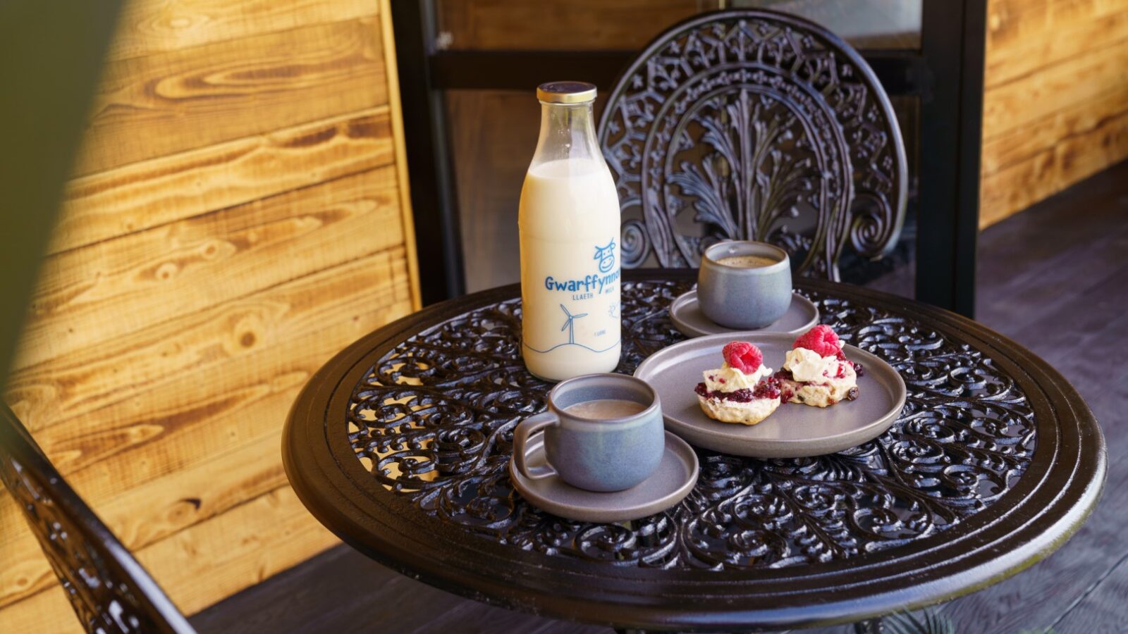 On a wooden patio sits a Piglet Pod beside bottled milk, two cups of coffee, and pastries with fruit, all elegantly arranged on a decorative metal table.