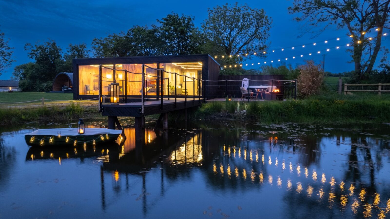 A modern glass cabin with lit string lights sits by a pond at dusk, reflecting in the water like a rustic boathouse. Trees and a fire pit are nearby, adding charm to the serene setting.