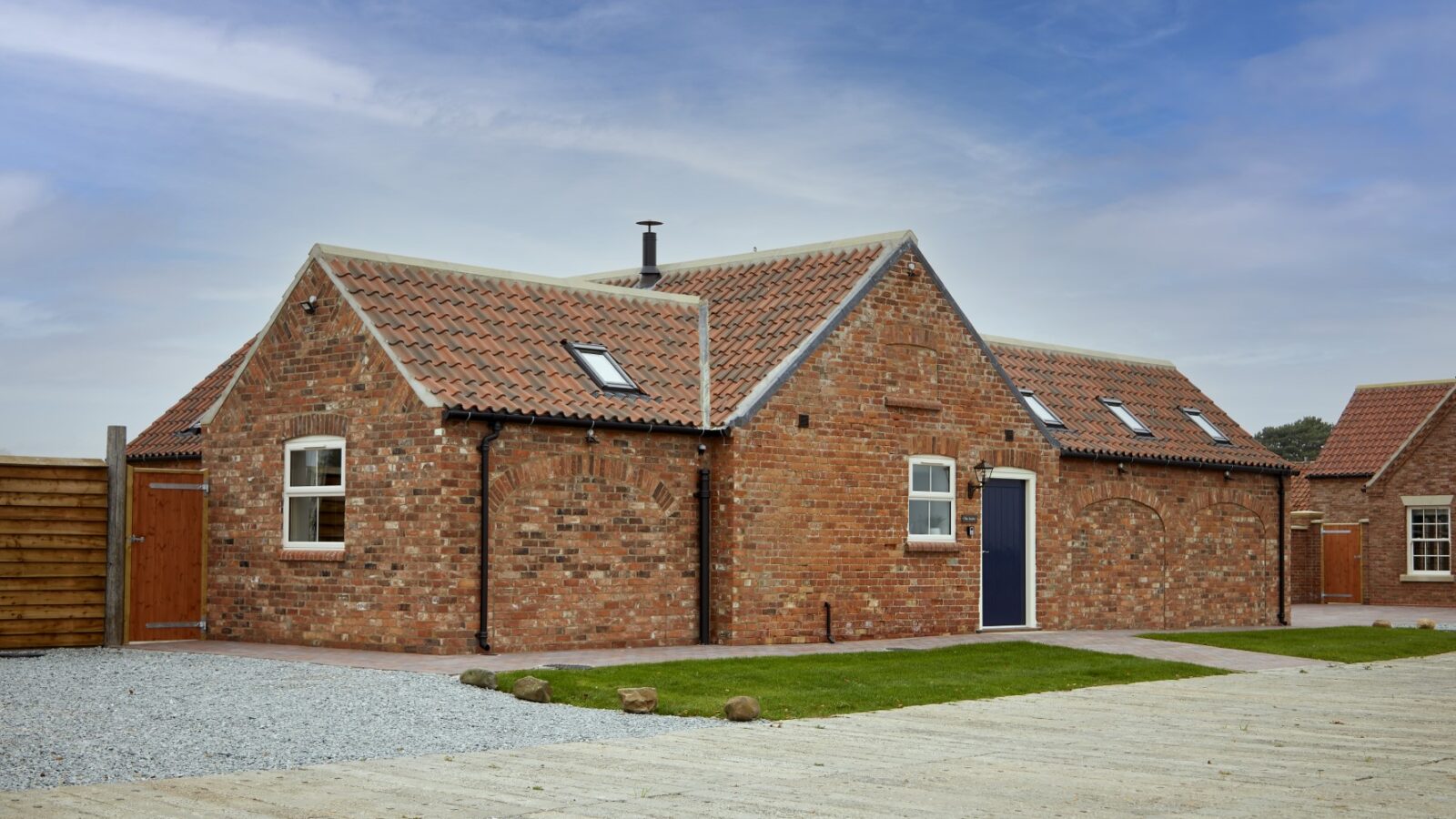 The Stables is a charming brick house with a red tile roof, multiple windows, and a dark blue door. It sits gracefully on a gravel path and grass lawn under a cloudy sky, offering proximity to exciting equestrian activities and horse riding opportunities.