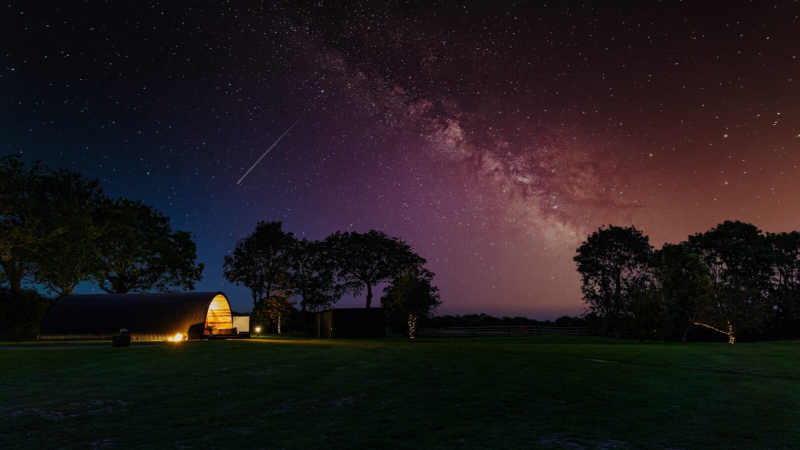 Beneath the starry night sky, a glowing Piglet Pod nestles amidst trees on a grassy field, with the Milky Way casting its ethereal light above.