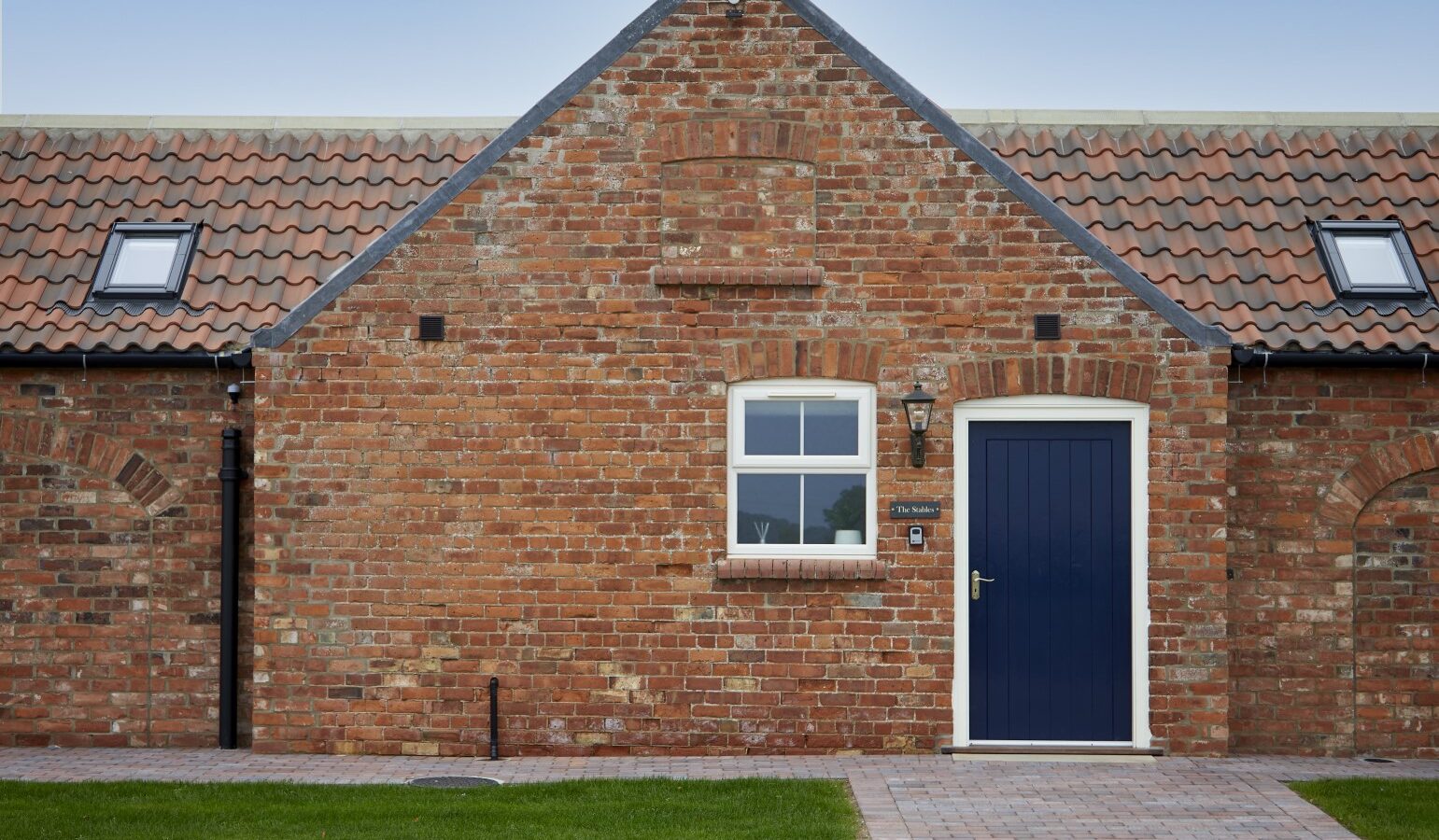 The charming red brick house with a blue door, small window, and sloped roof sits just beyond the stables. A brick pathway leads to the entrance, echoing the elegance of nearby equestrian activities.