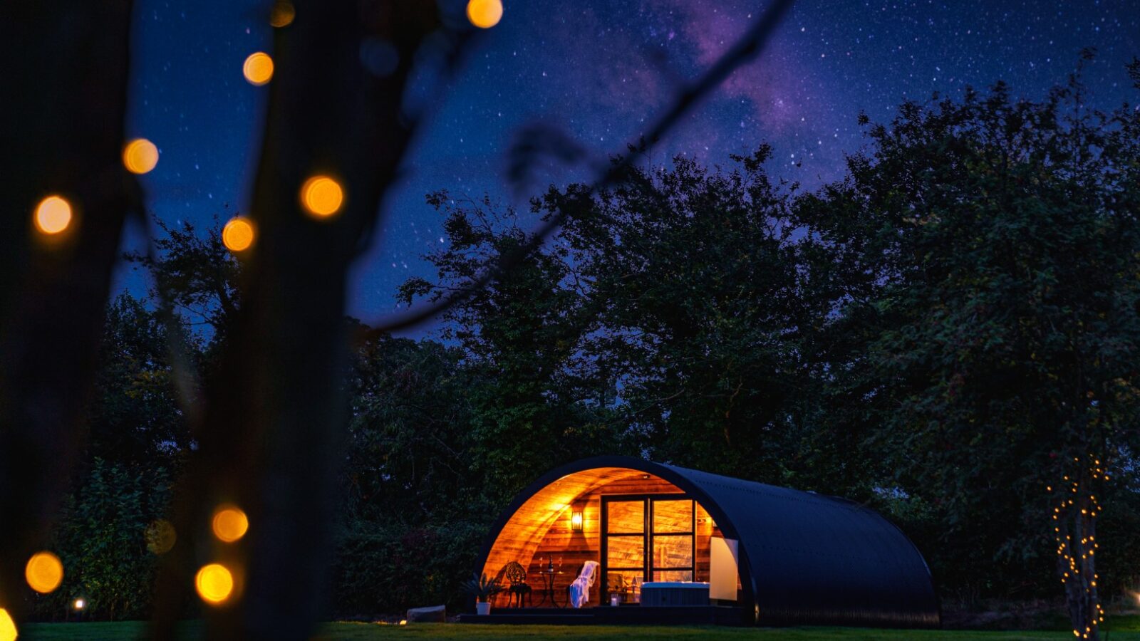 Nestled among the trees, the Piglet Pod offers a modern cabin experience with warm interior lights illuminating against a starry night sky, while small outdoor lights add a magical touch to the serene setting.