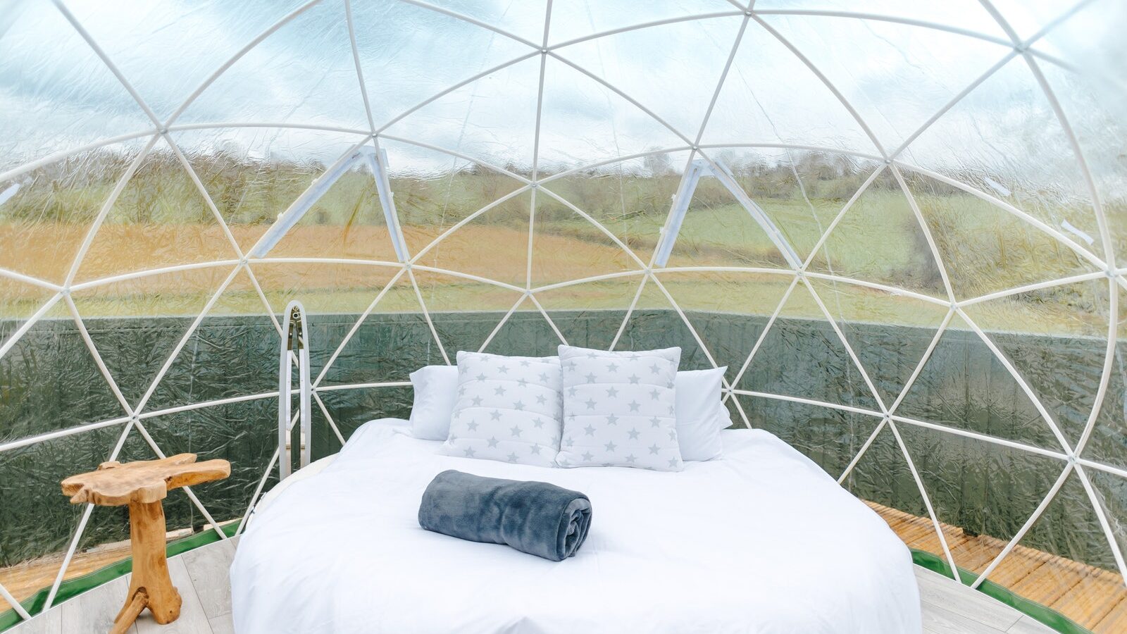 A clear geodesic dome at Chicken Shed Lodge features a white bed, plush pillows, and a neatly folded blanket, all nestled against a lush grassy landscape. A small wooden table stands charmingly beside it.