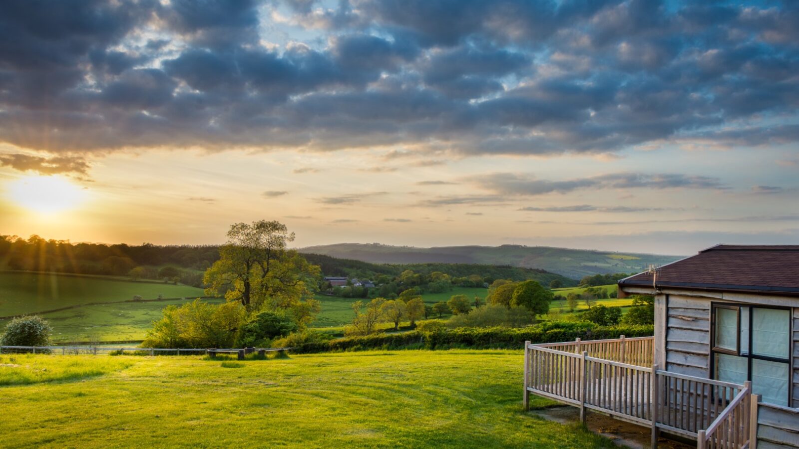 Sunset over a green countryside landscape with rolling hills and Moonrise Lodges nestled on the right under a partly cloudy sky.