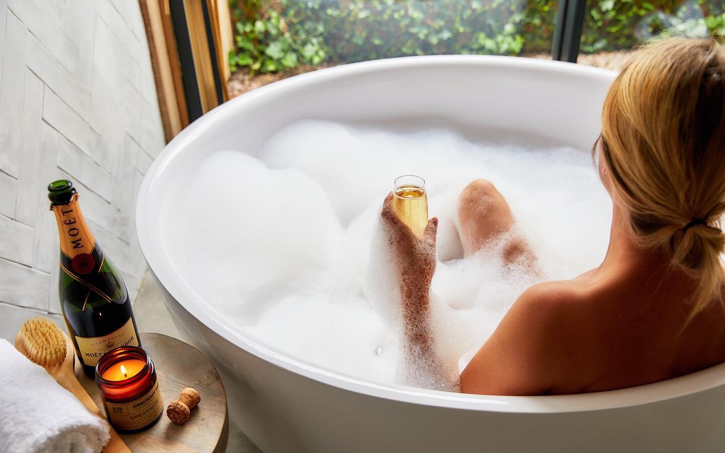 A person relaxing in a bubble bath, holding a glass of Moët champagne, enjoys the tranquil atmosphere. Nearby, a lit Dolassey candle casts a warm glow over the scene as lush greenery can be seen through the window. A bath brush rests invitingly beside the bottle of champagne.