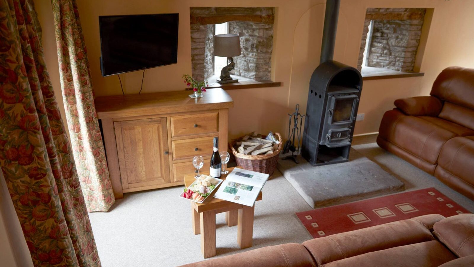 The Upper Barn's cozy living room features leather couches, a wood stove, and a TV. A small table with wine, glasses, and a book adds charm to the inviting space.