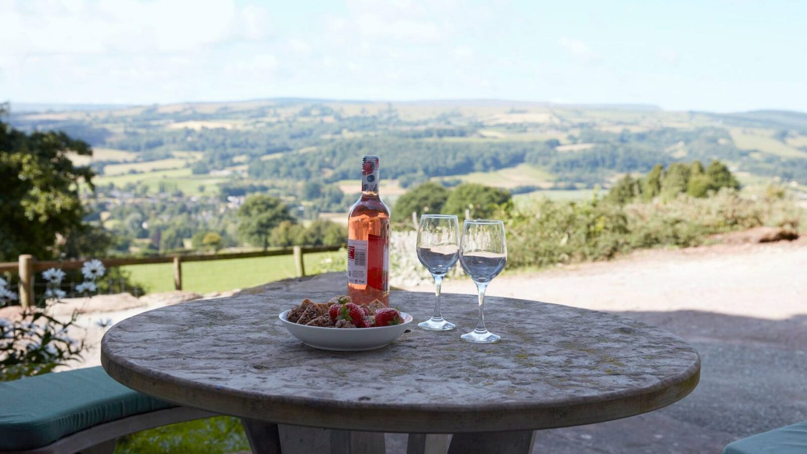 A stone table at Upper Barn, adorned with wine, strawberries, and glasses, overlooks a scenic countryside landscape under a clear sky.