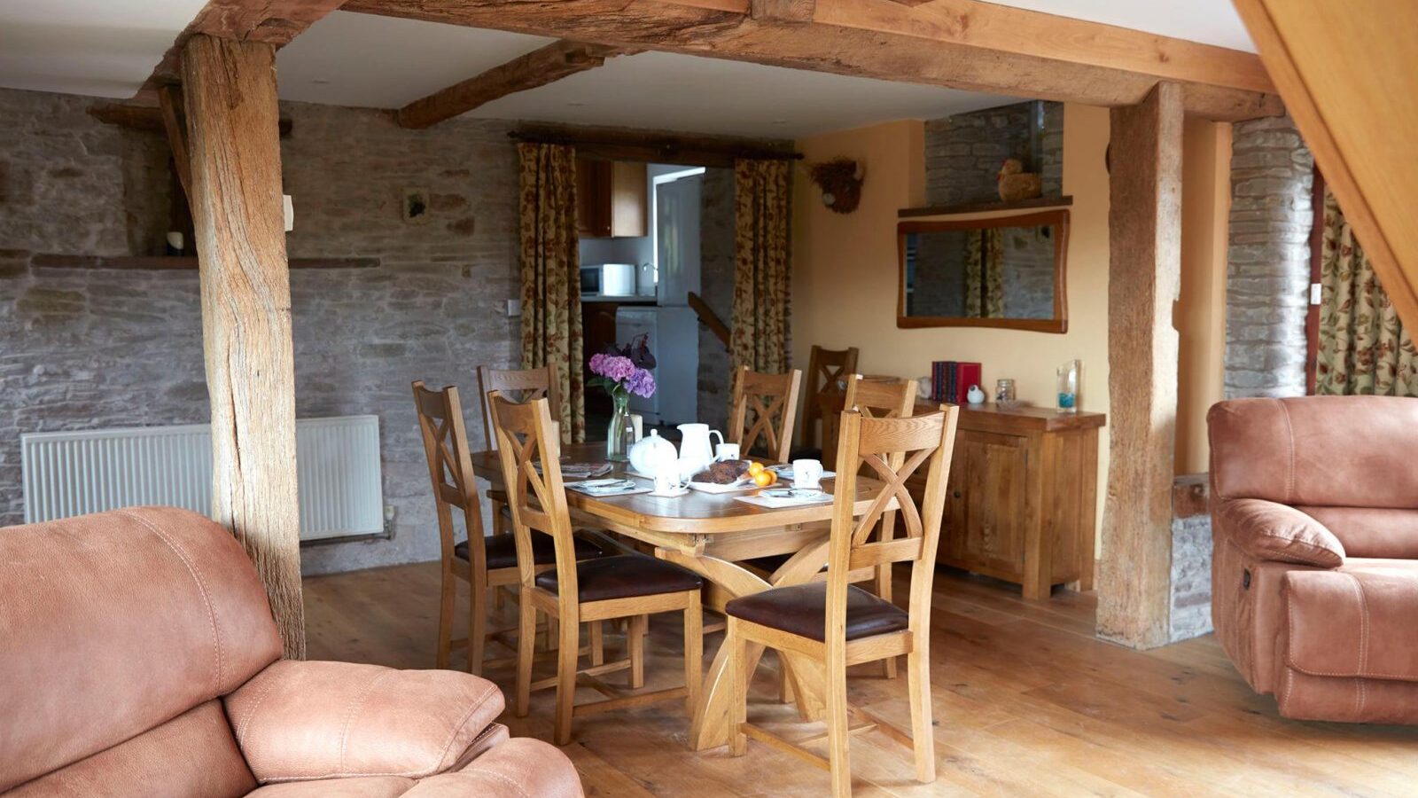 Upper Barn's rustic dining room features wooden beams, a table set for breakfast, plush sofas, and inviting stone walls.