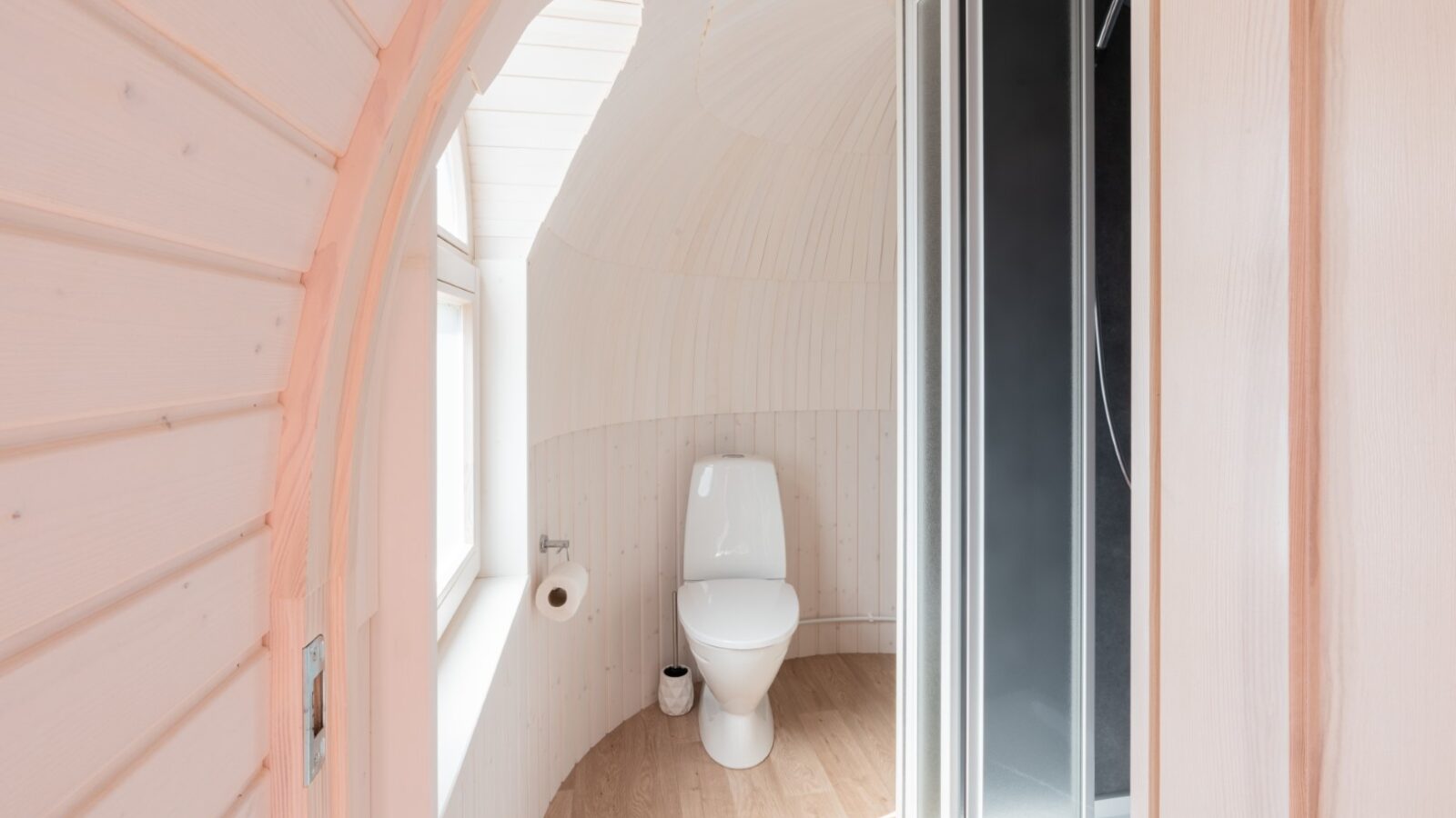 In this white bathroom with a curved wooden wall, where the toilet and holder sit neatly alongside a rock-inspired, black-framed glass shower, you'll find an inviting escape that balances rustic charm and modern luxury.