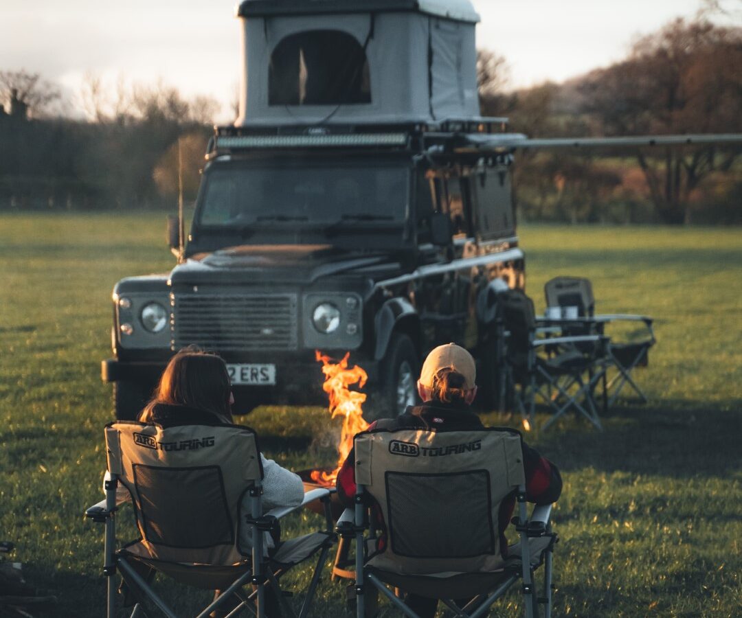 Two people sit in camping chairs by a firepit near a Defender with a rooftop tent, set in a grassy area at dusk, enjoying the serenity of camping.