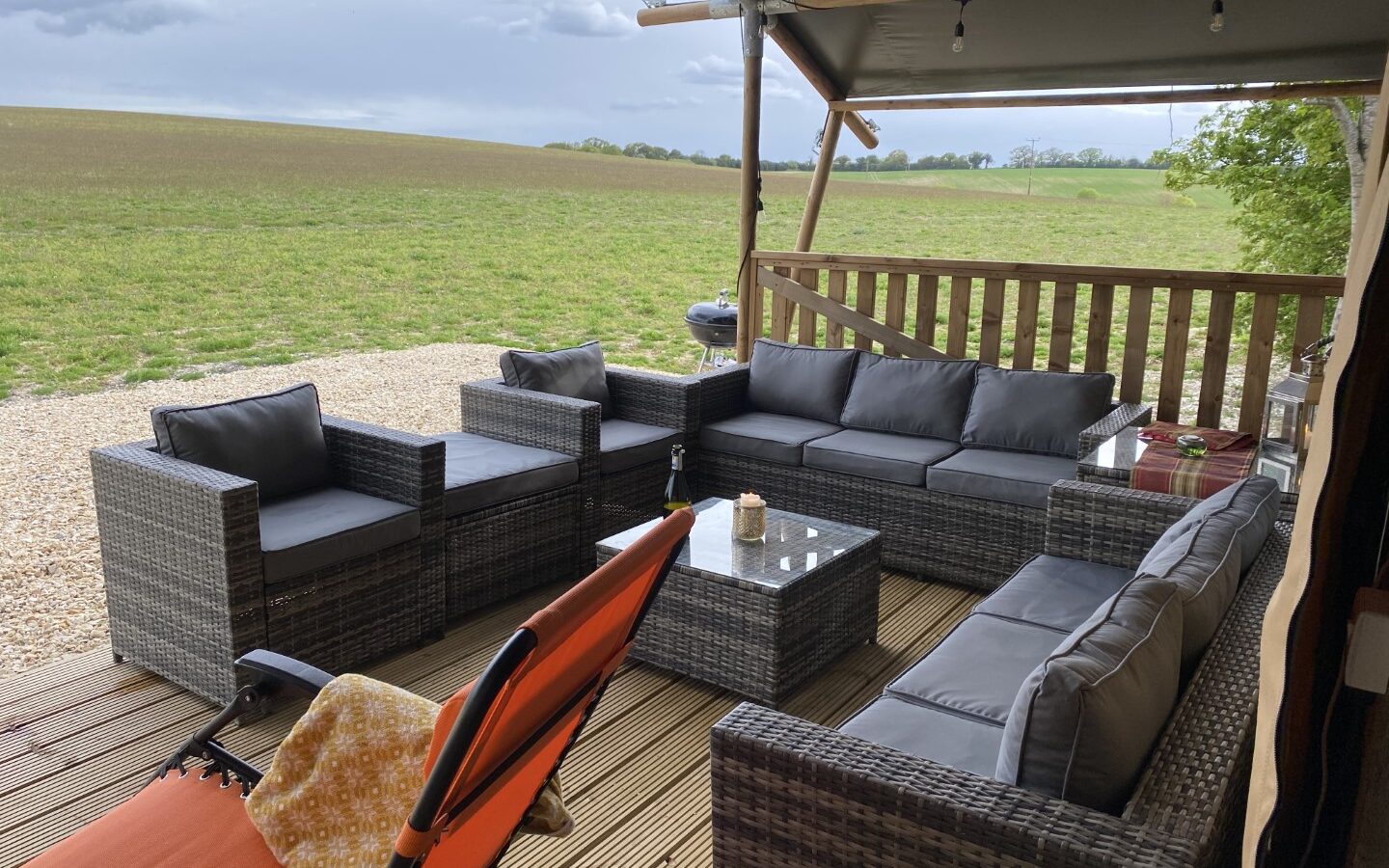 Outdoor patio with gray wicker furniture set, orange chair, and glass table, offering a glamping vibe with a view of a cloudy sky over a grassy field reminiscent of the Chilterns View.