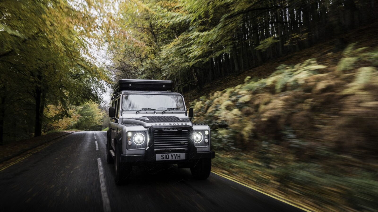 A Land Rover Defender navigates a forested area on a damp road, surrounded by green and brown foliage, perfect for a camping adventure.