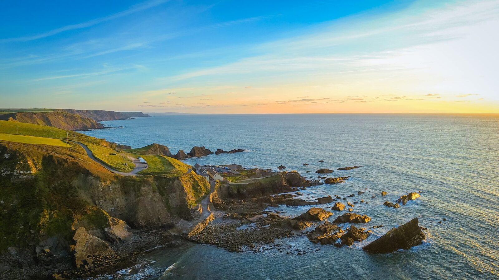 Aerial view of a Devon coastal landscape at sunset, featuring rugged cliffs and rolling green hills. The sky is a gradient of blue and orange, casting a warm glow over the scene. Waves crash gently against the rocks below, reminiscent of tales from the Sleepy Owl nearby.