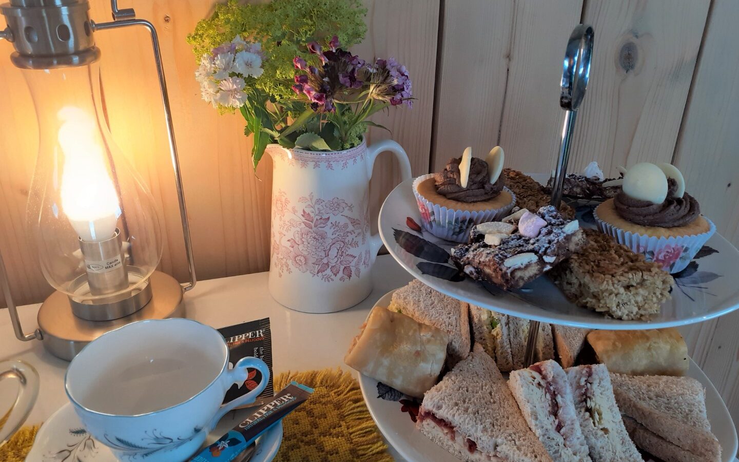 A tiered stand with sandwiches, pastries, and cupcakes sits beside tea cups, a teapot, and a floral vase on a quaint table in the Shepherds Hut at Cae Main. Softly lit by a nearby lamp, it offers an inviting accommodation for anyone seeking cozy afternoon indulgence.