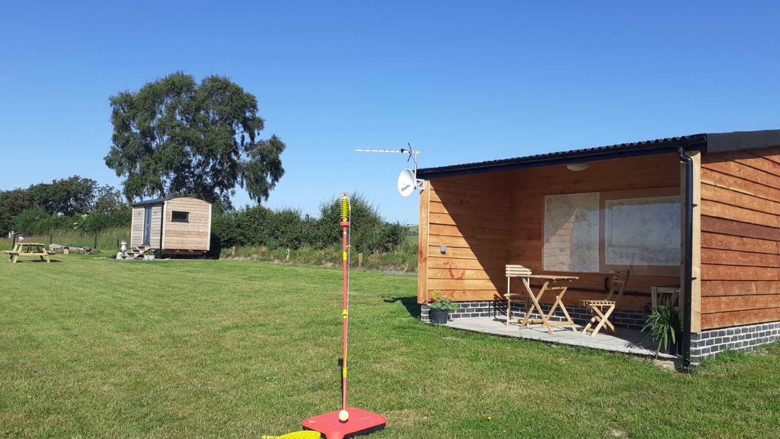 A charming shepherd's hut sits beside a wooden cabin with an outdoor seating area next to a grassy field. A tetherball set stands under the clear blue sky, offering a delightful scene at Cae Main.