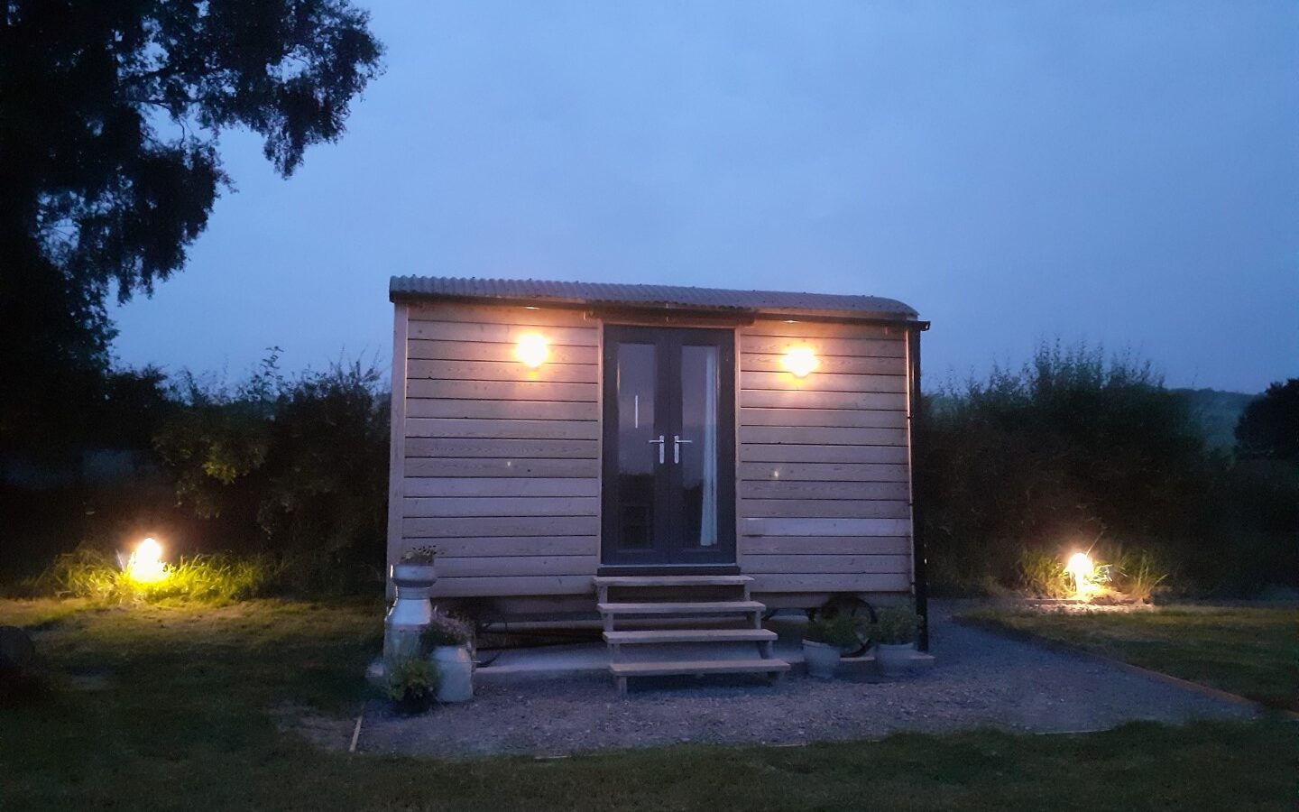 At dusk, a small wooden cabin resembling a charming shepherd's hut is illuminated by exterior lights. Surrounded by lush grass and tall trees, the Cae Main retreat invites you in, with its gently worn steps leading to an inviting door.