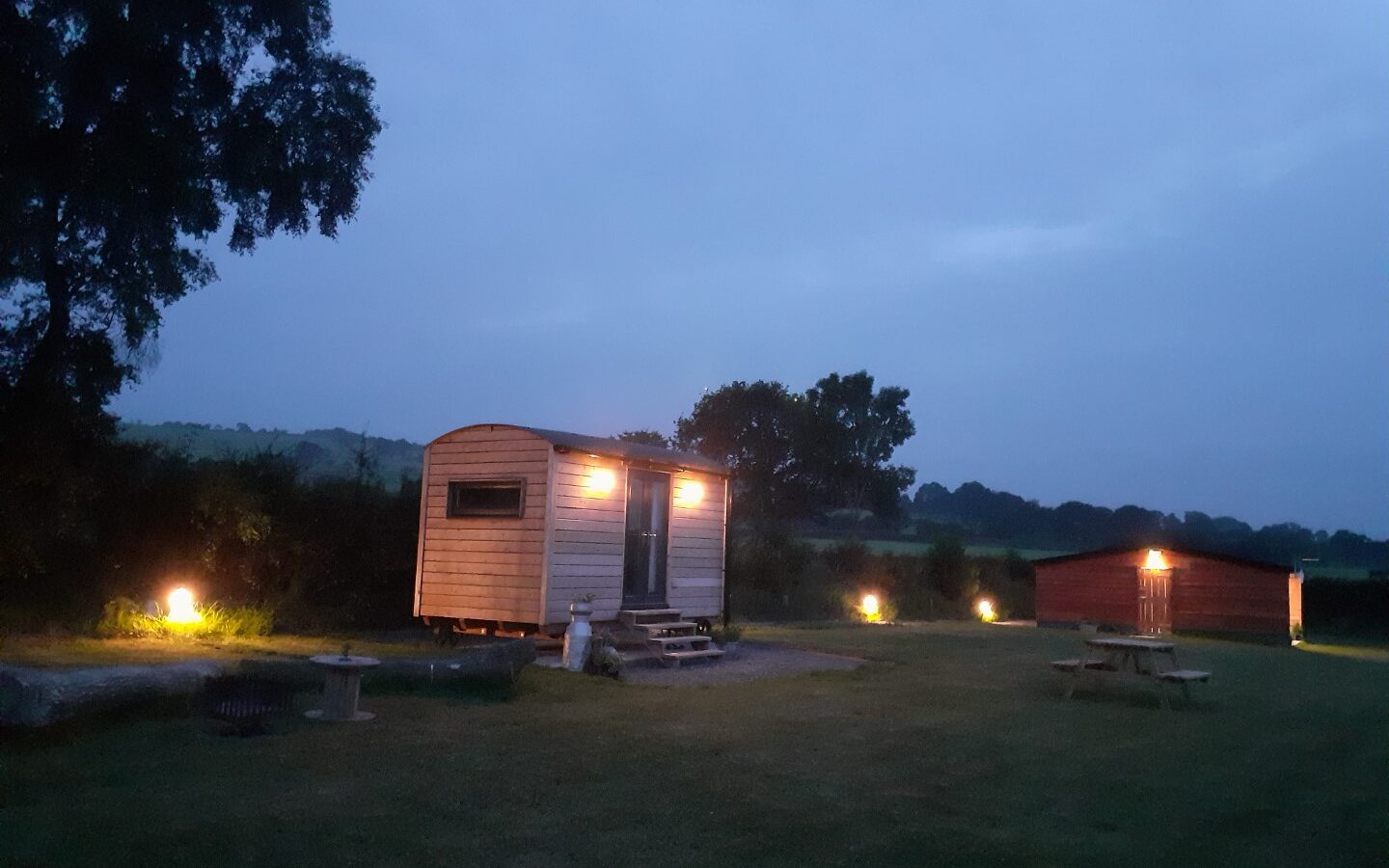 Nestled in a grassy field with distant hills, Cae Main offers a charming accommodation: a small wooden cabin glowing at dusk. Enjoy the tranquil setting with picnic tables scattered around this cozy shepherd's hut, perfect for an idyllic getaway.