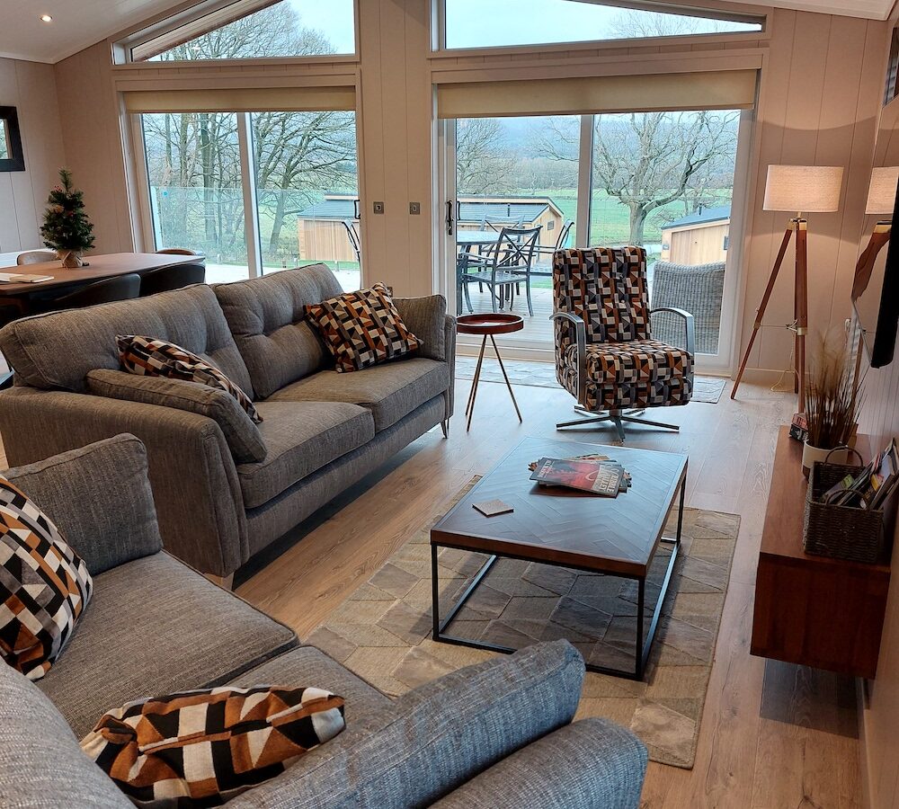 A modern living room in Bowland Retreat features gray sofas, patterned cushions, a coffee table, and large windows overlooking a patio and lush landscape, offering the perfect blend of comfort and nature, reminiscent of quaint lodges.