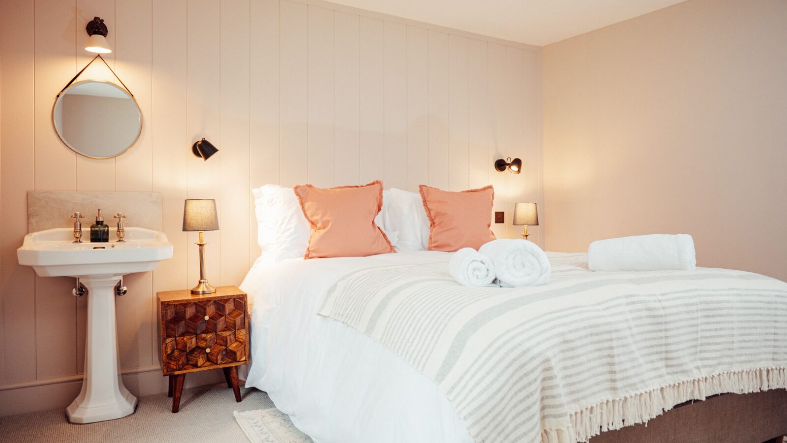 The cozy Almer Manor bedroom features a double bed adorned with white bedding and peach pillows, complemented by elegant wall lamps. A round mirror hangs above a quaint sink with a cabinet, enhancing the room's inviting charm.