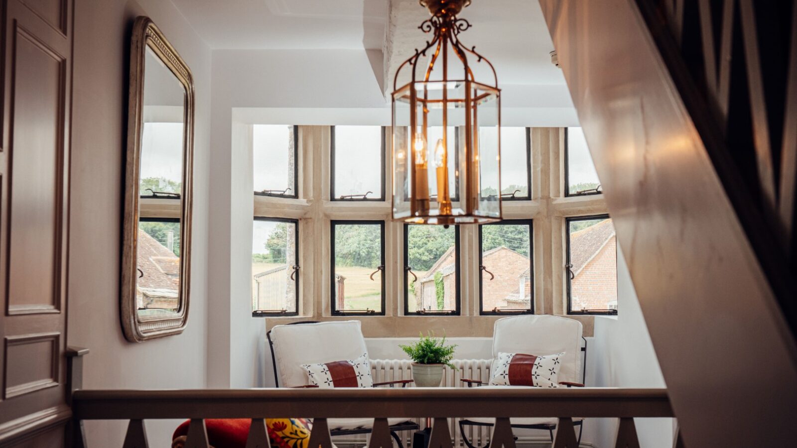 The Almer Manor-inspired cozy seating area features chairs, cushions, and a small plant by a bay window, complemented by a chandelier hanging from the ceiling with an inviting outdoor view.