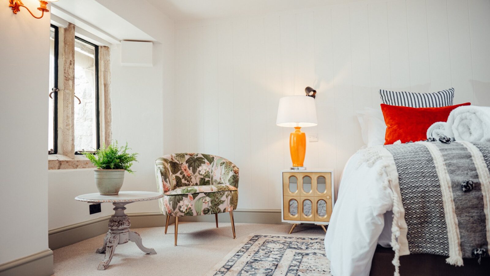 Experience the charm of Almer Manor in this cozy bedroom featuring a plush bed, a floral chair, and a classic wooden cabinet. A round table with a plant sits by the window, all beautifully illuminated by soft lamp lighting.