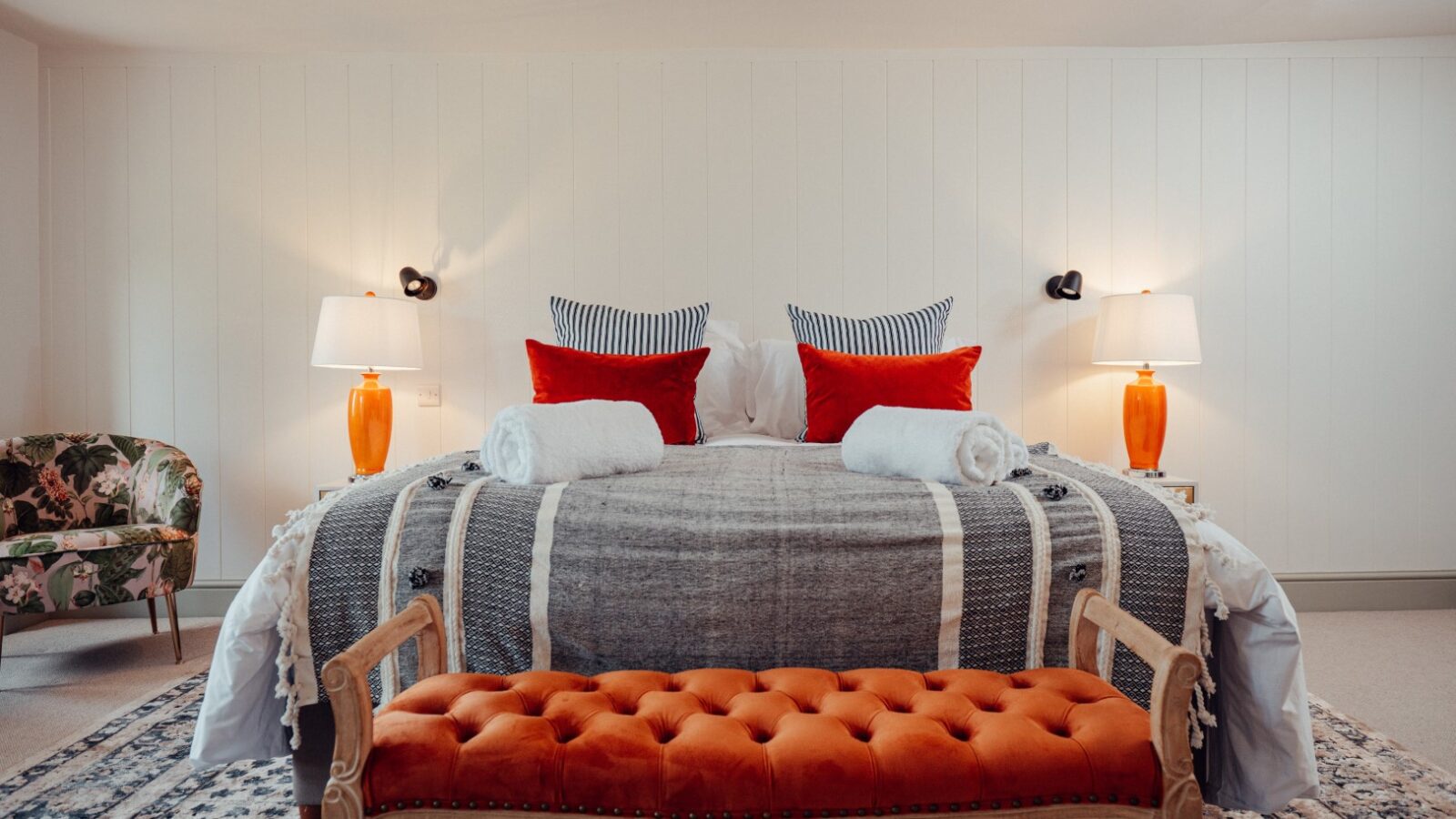 The elegant bedroom at Almer Manor features a double bed adorned with vibrant orange pillows and lamps, complemented by a wooden bench with orange upholstery and a stylish patterned armchair.