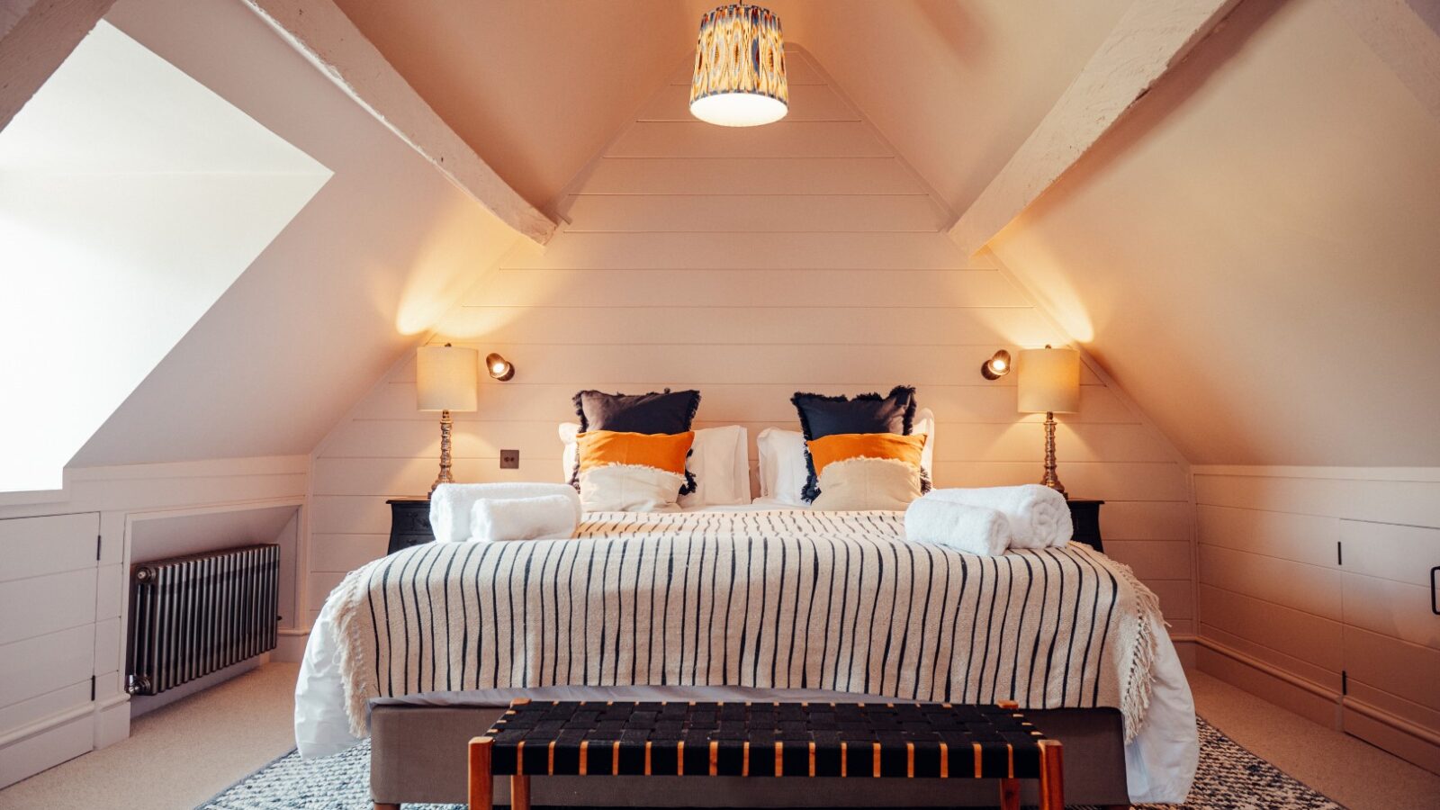 The cozy attic bedroom at Almer Manor features a striped bedspread, plush pillows, two ambient lamps, and a bench at the foot of the bed.