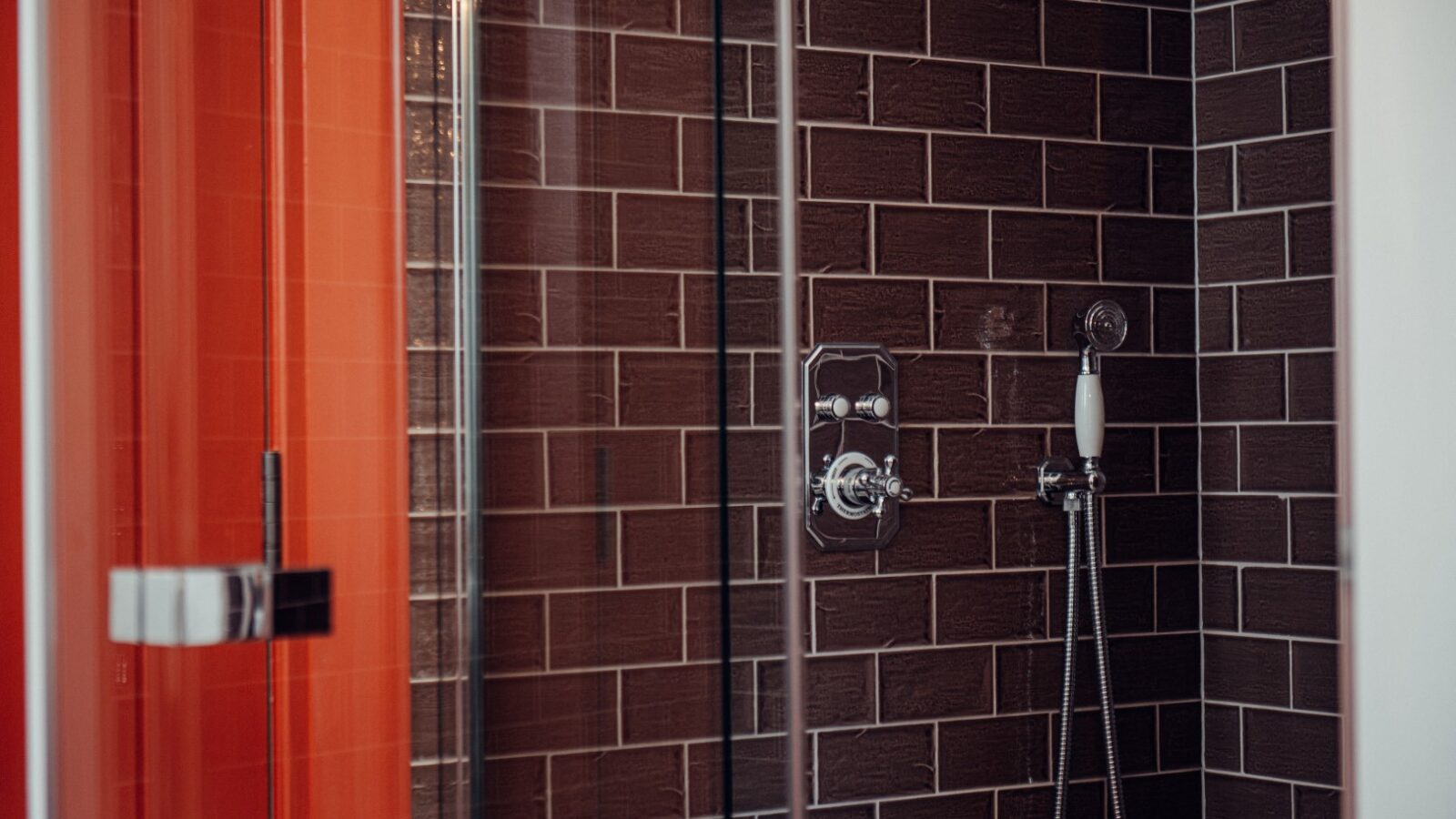 The Almer Manor shower features sleek dark brown subway tiles, a glass door, and a handheld showerhead with polished chrome fixtures, adding a touch of elegance to any bathroom.