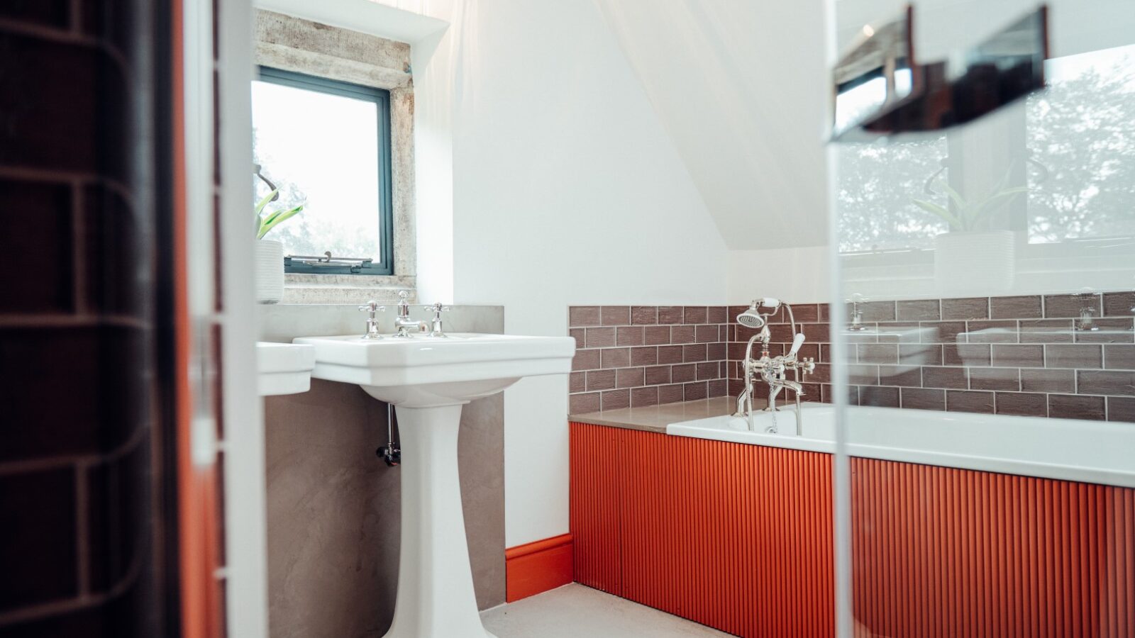 The bathroom at Almer Manor features a white pedestal sink and an orange bathtub, complemented by brown tile accents. A window above the sink adds natural light to the charming space.