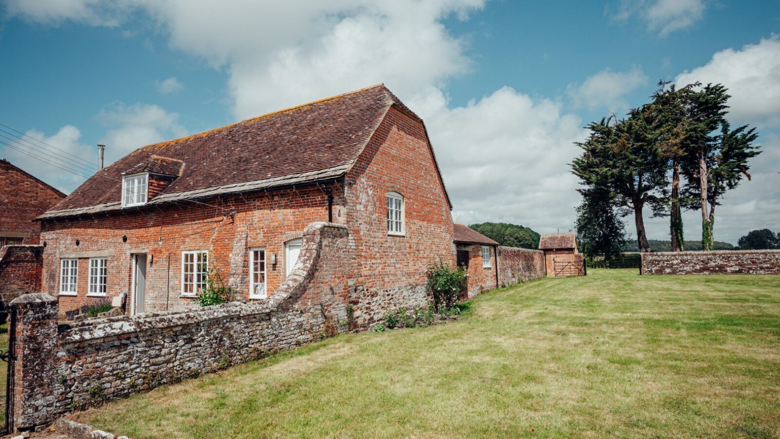 A brick house with a sloped roof and stone wall in the grassy yard of Almer Manor, nestled under a partly cloudy sky.